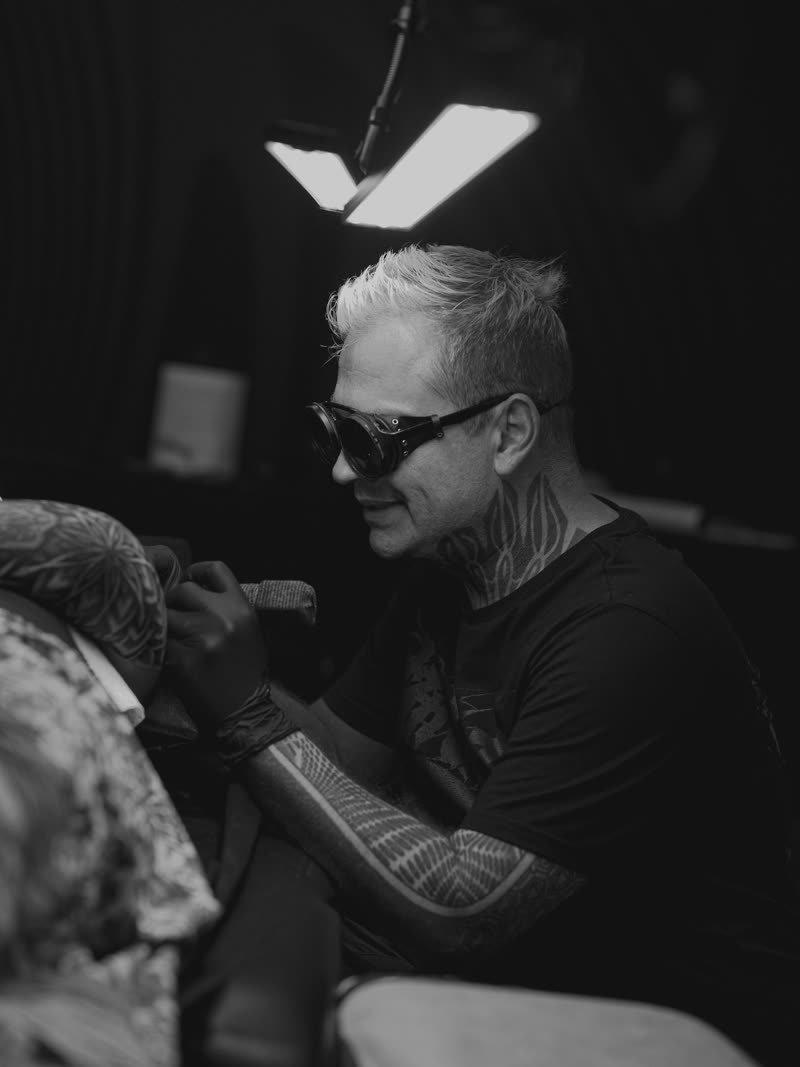 Ilya Cascad tattooing in sunglasses under a single hard light, black and white