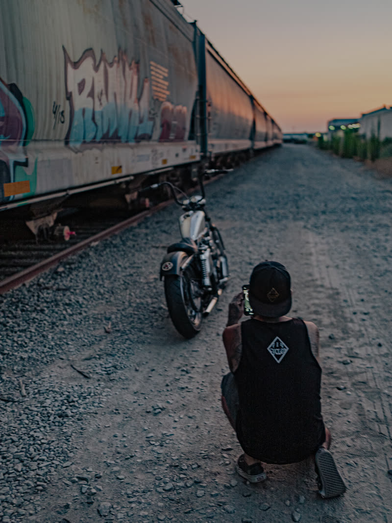 Rider crouched on the gravel beside a chopper, freight cars running to the sunset
