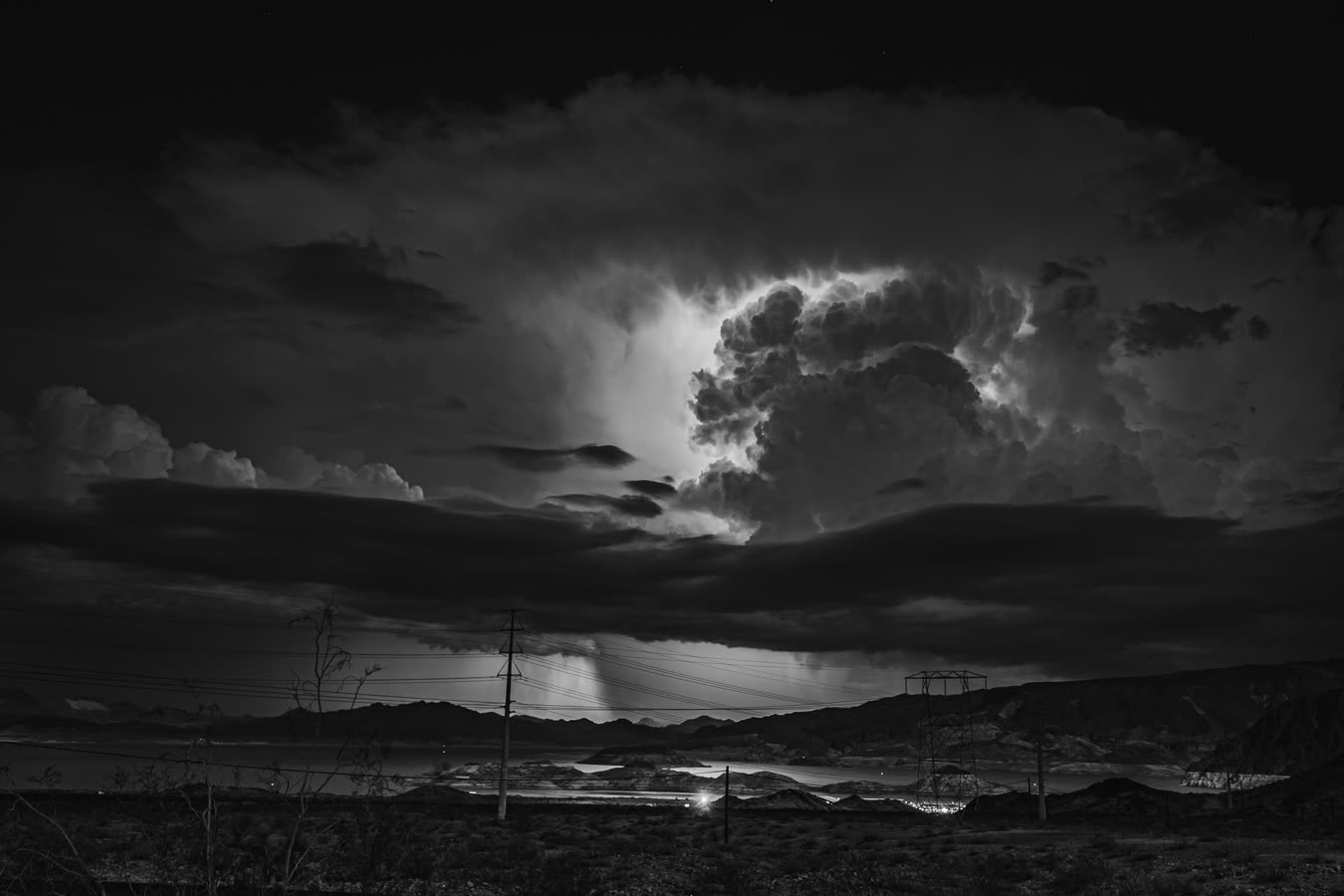 A cumulonimbus lit from inside above transmission towers and the lake